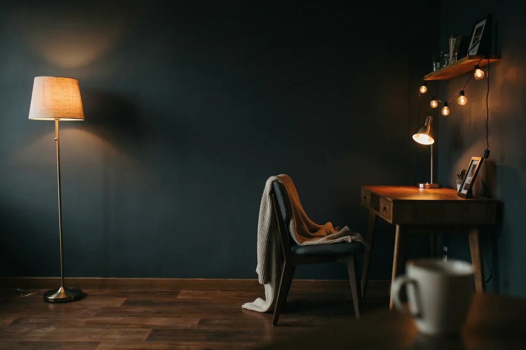 Warm-lit room corner showing light at three heights, a floor lamp low in the corner, a small desk lamp on a wooden desk and warm bulbs along a shelf, with the ceiling light off
