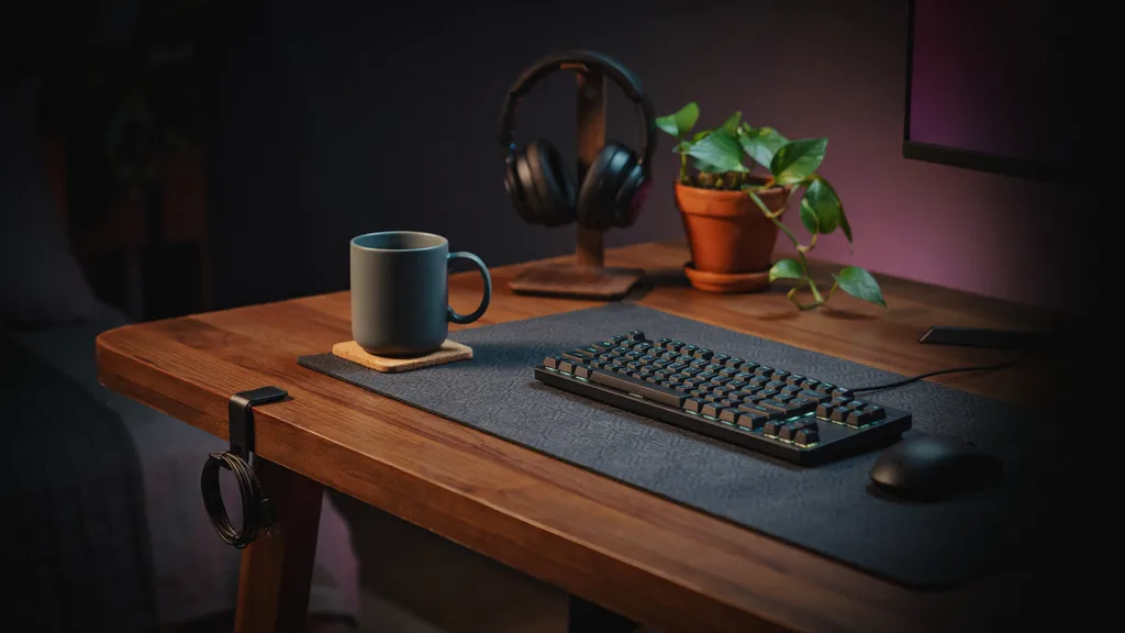 Warm nighttime gaming desk at desk level with a large patterned desk mat, low-profile keyboard, a mug resting on a cork-backed coaster and cables clipped to the desk edge