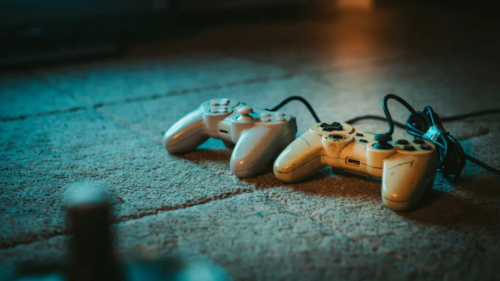 Two mismatched wired game controllers side by side on a worn carpet, one clean and pale, one yellowed with a taped cable, lit by soft television glow