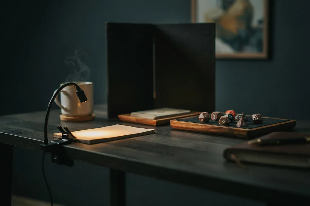 The game master's end of a tabletop RPG table with a plain folding screen, a felt lined wooden dice tray of polyhedral dice, a clip lamp angled onto blank notes and a mug set well away from the paper