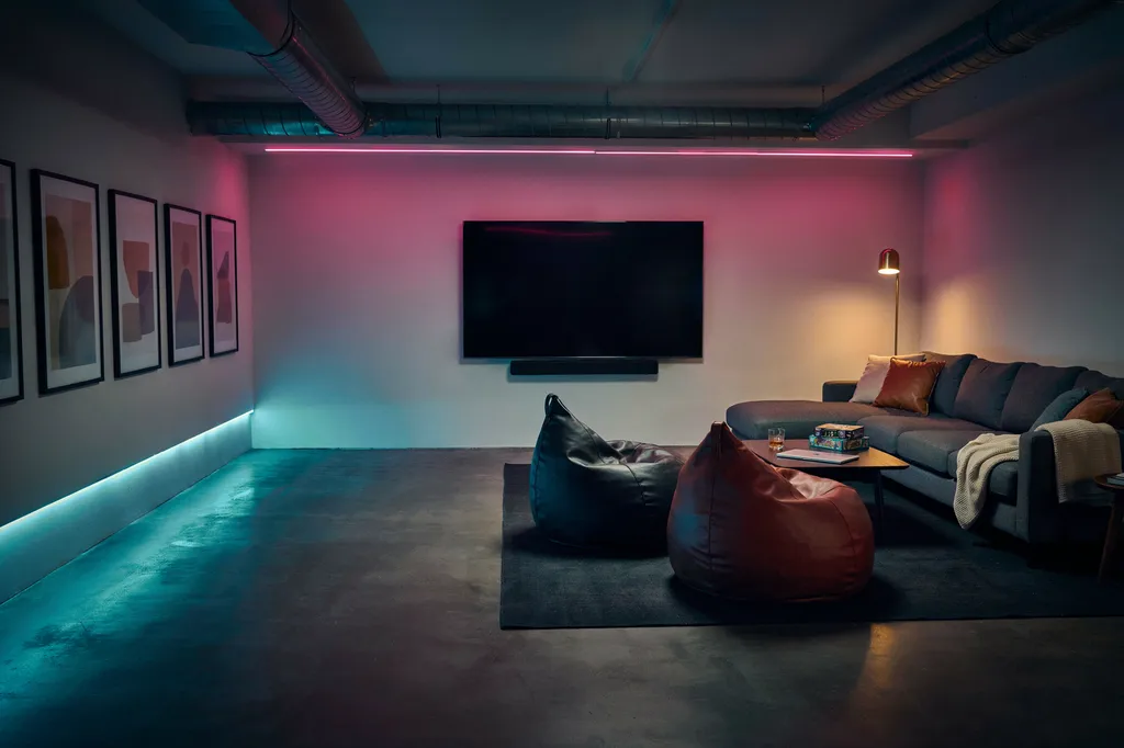 Basement gaming lounge with a wall-mounted screen, sectional couch, beanbags, magenta and teal accent lighting and a row of framed prints along the side wall