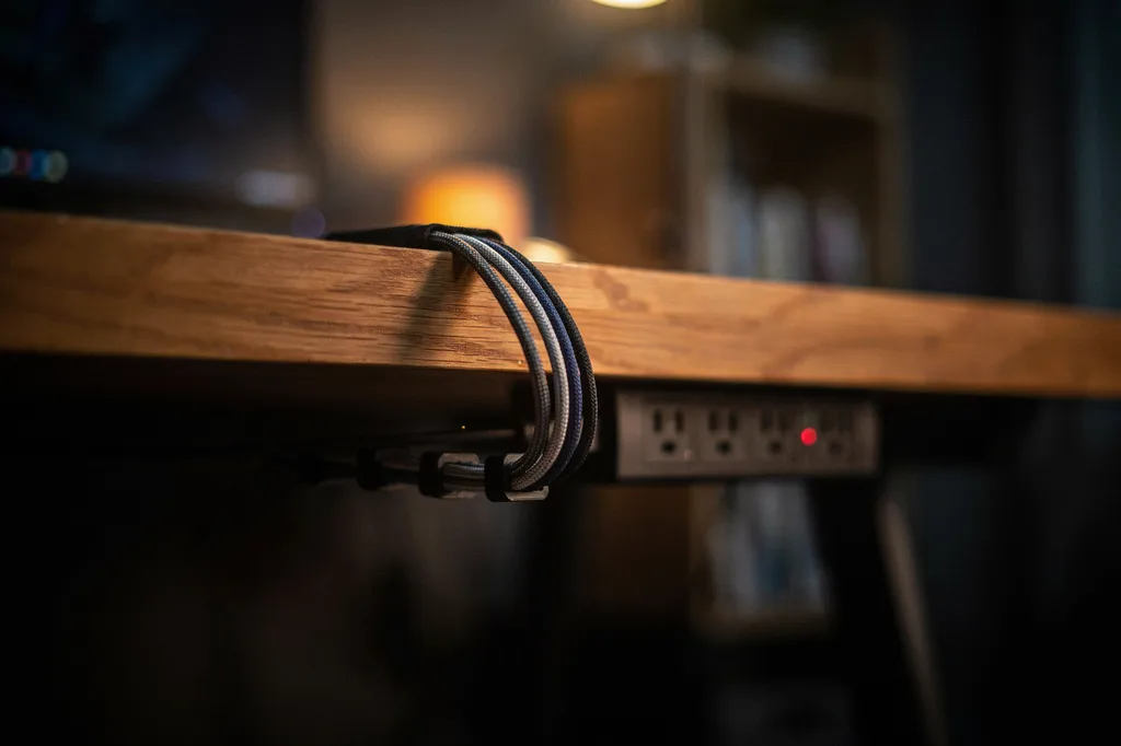 Cable management under the back edge of a wooden gaming desk, with braided cables in a fabric sleeve held by clips running down the desk leg