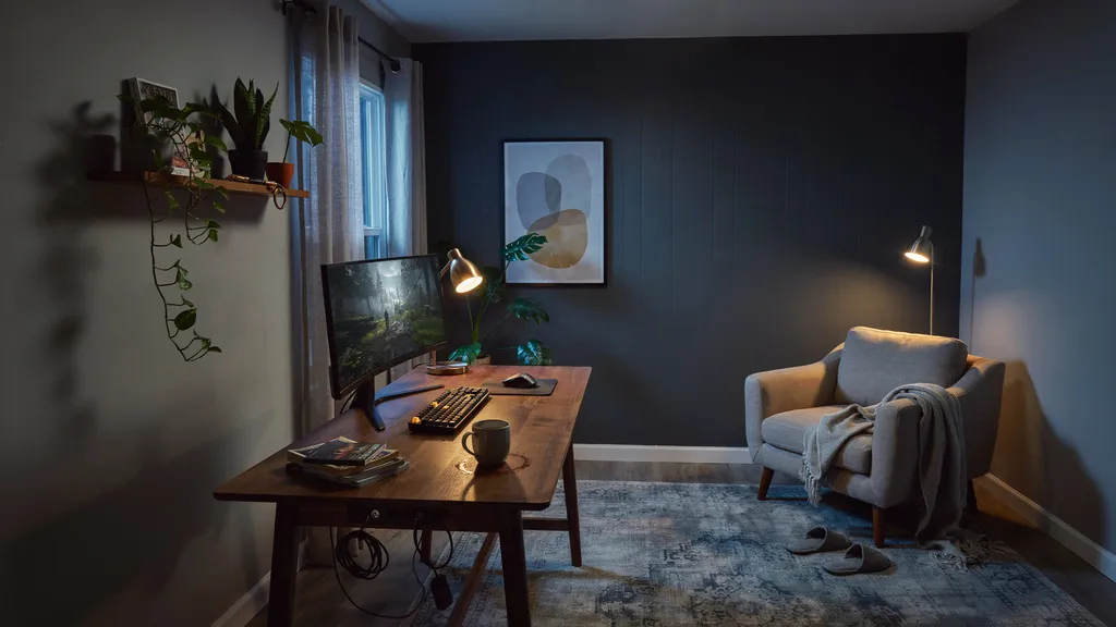 Gaming room with a dark charcoal accent wall, one large framed art print above the desk, warm layered lamp light and a reading chair in the corner