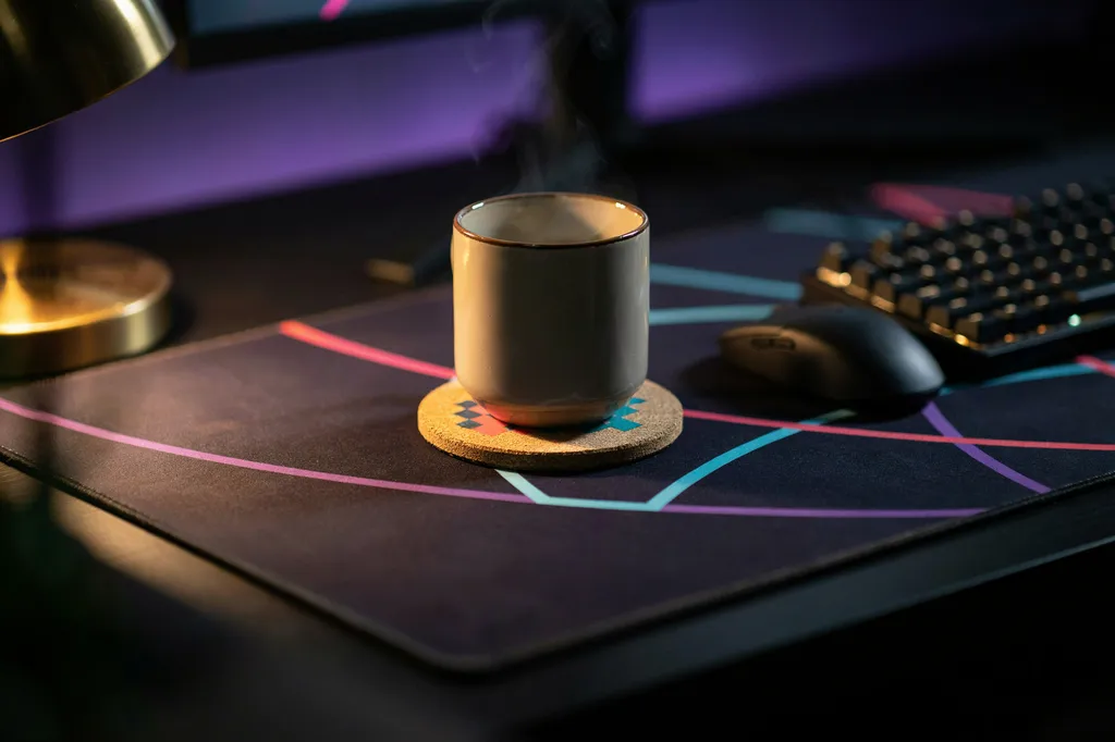 Close-up of a small gaming desk with a patterned desk mat, a coaster and a warm mug under lamp light