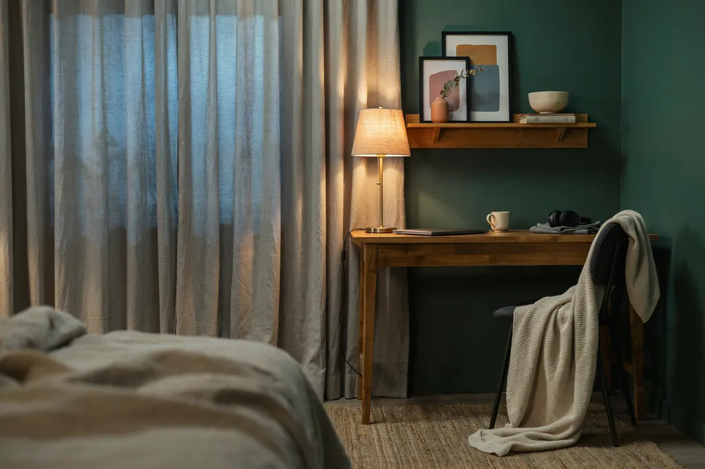 Cozy bedroom gaming corner with warm amber lamp light, deep green wall, three framed prints above an oak desk and a knitted blanket on the chair